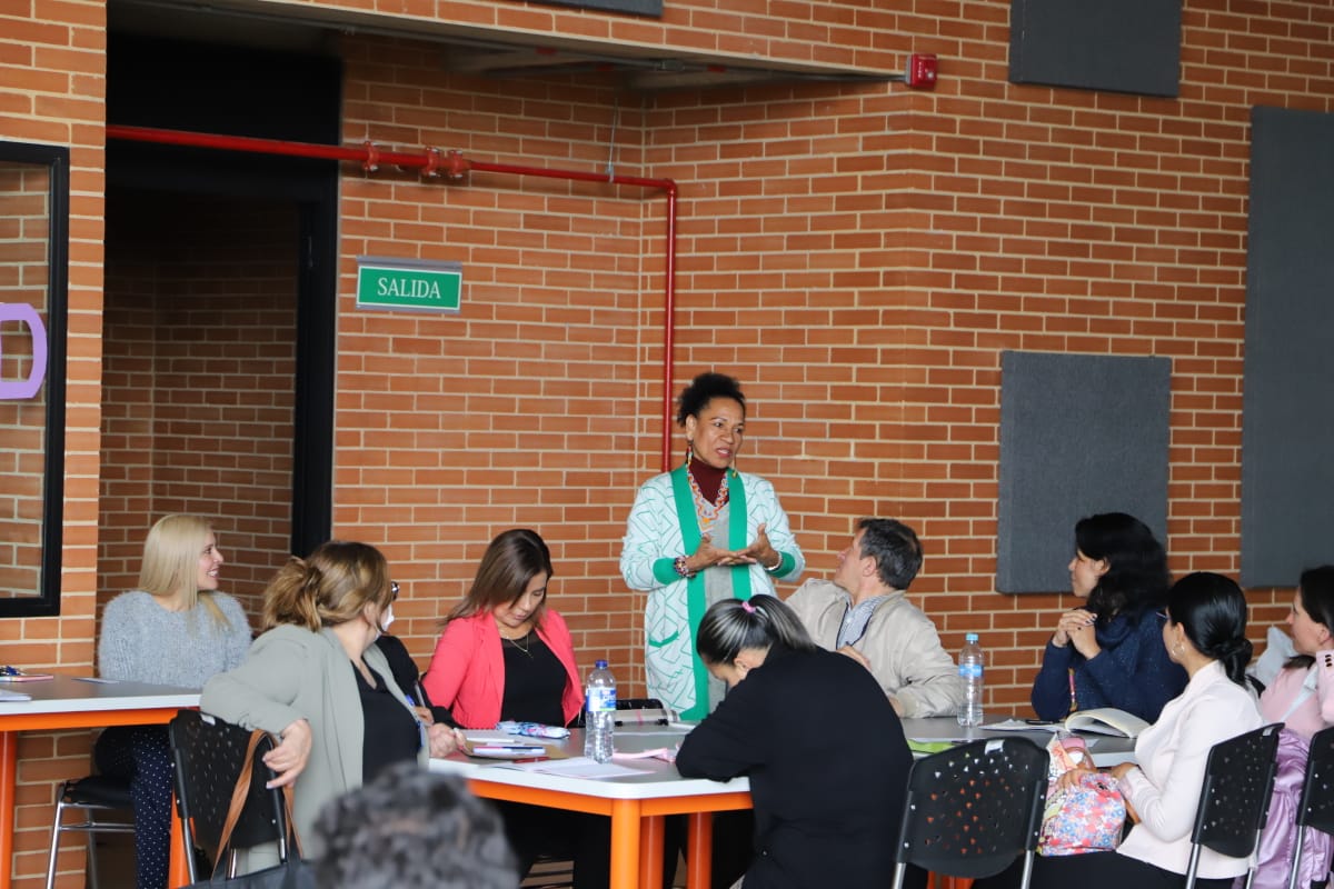 Profesora hablando a varios asistentes sentados alrededor de una mesa